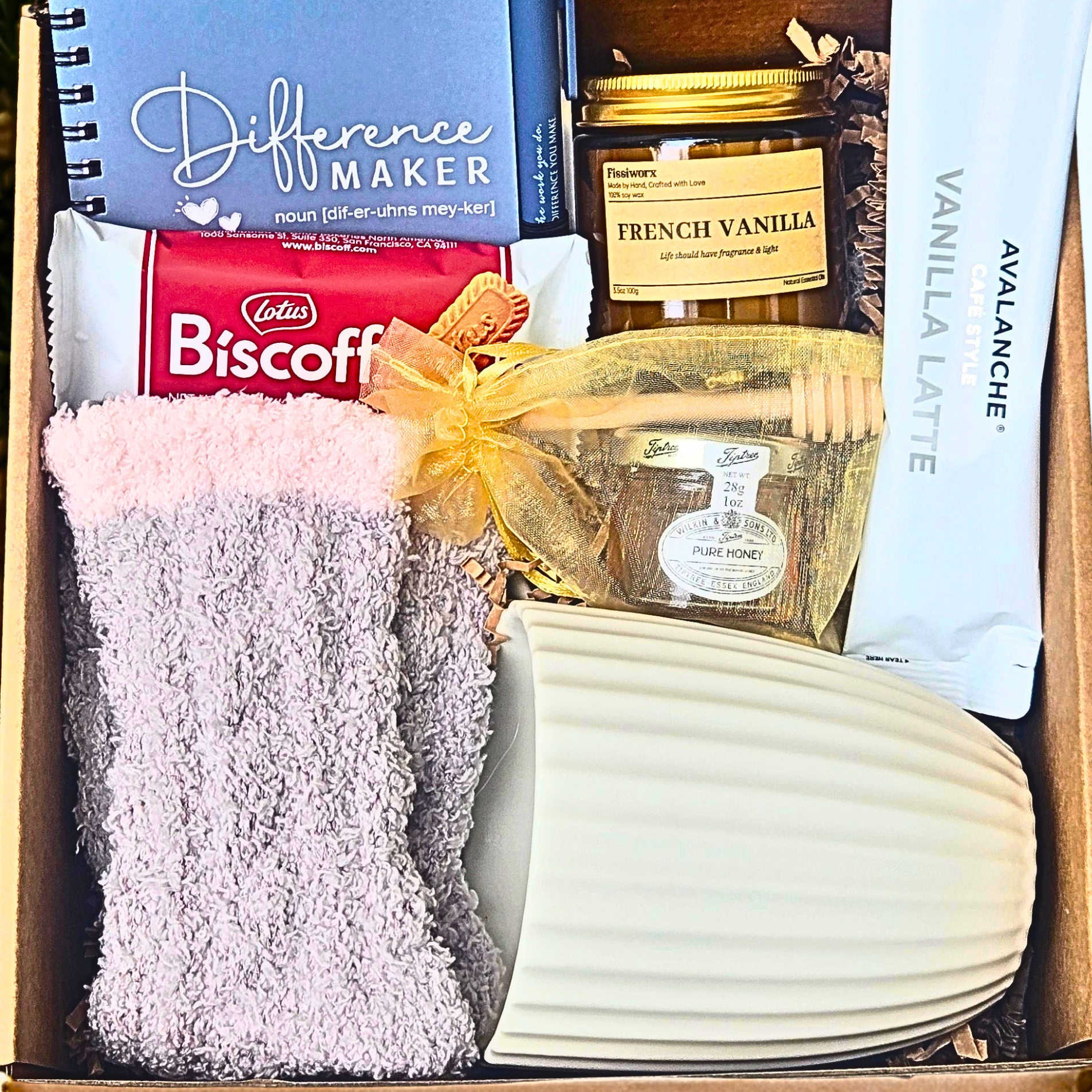 A curated gift box filled with assorted items including a blue Difference Maker notebook, a french vanilla candle, honey with dipper, a pack of Avalanche Vanilla Latte, fuzzy socks, a white ribbed mug and Biscoff cookies.