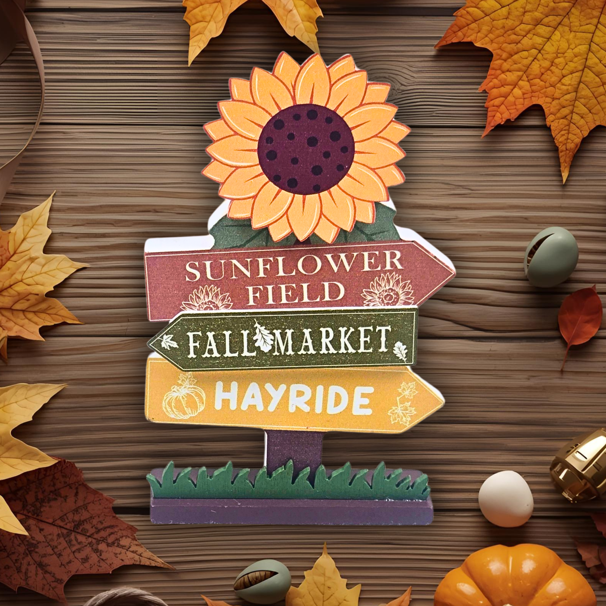 Decorative sign with sunflower design and autumn-themed text on a wooden surface with leaves and pumpkins.