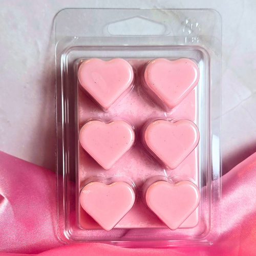 Pink heart-shaped wax melts in a clear plastic packaging on a pink fabric background