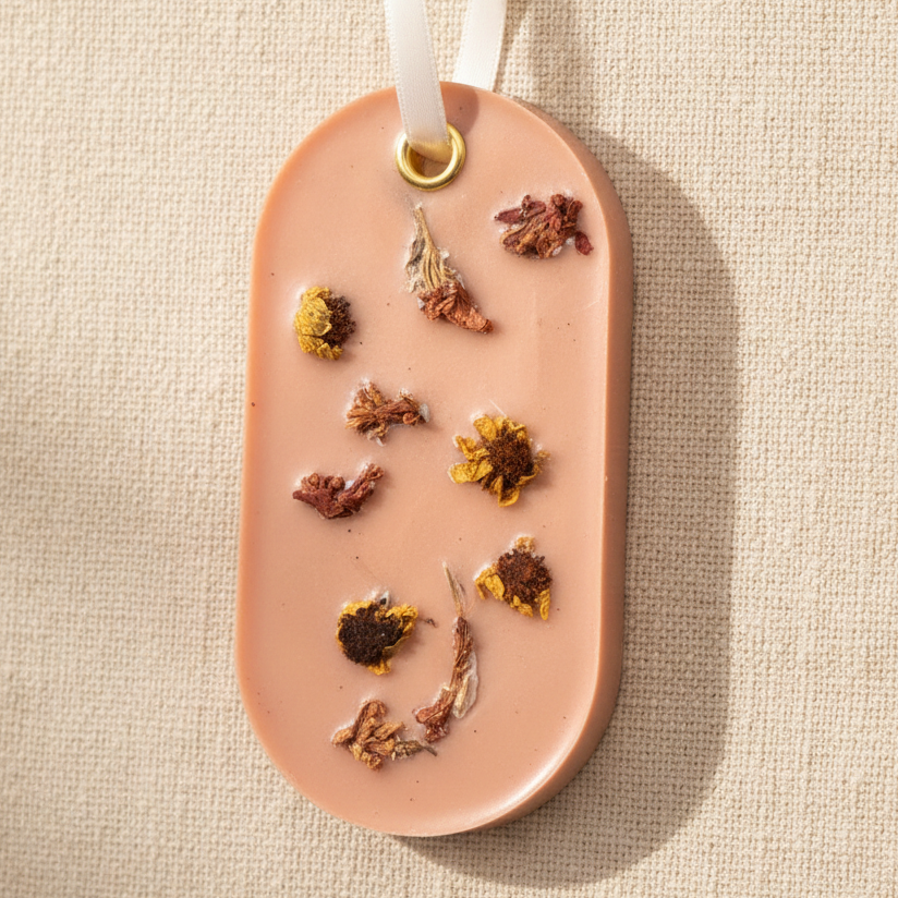 An oval light orange wax d with dried flower inclusions, hanging from a white ribbon with a gold ring, set against a textured beige linen background.