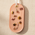 An oval light orange wax d with dried flower inclusions, hanging from a white ribbon with a gold ring, set against a textured beige linen background.