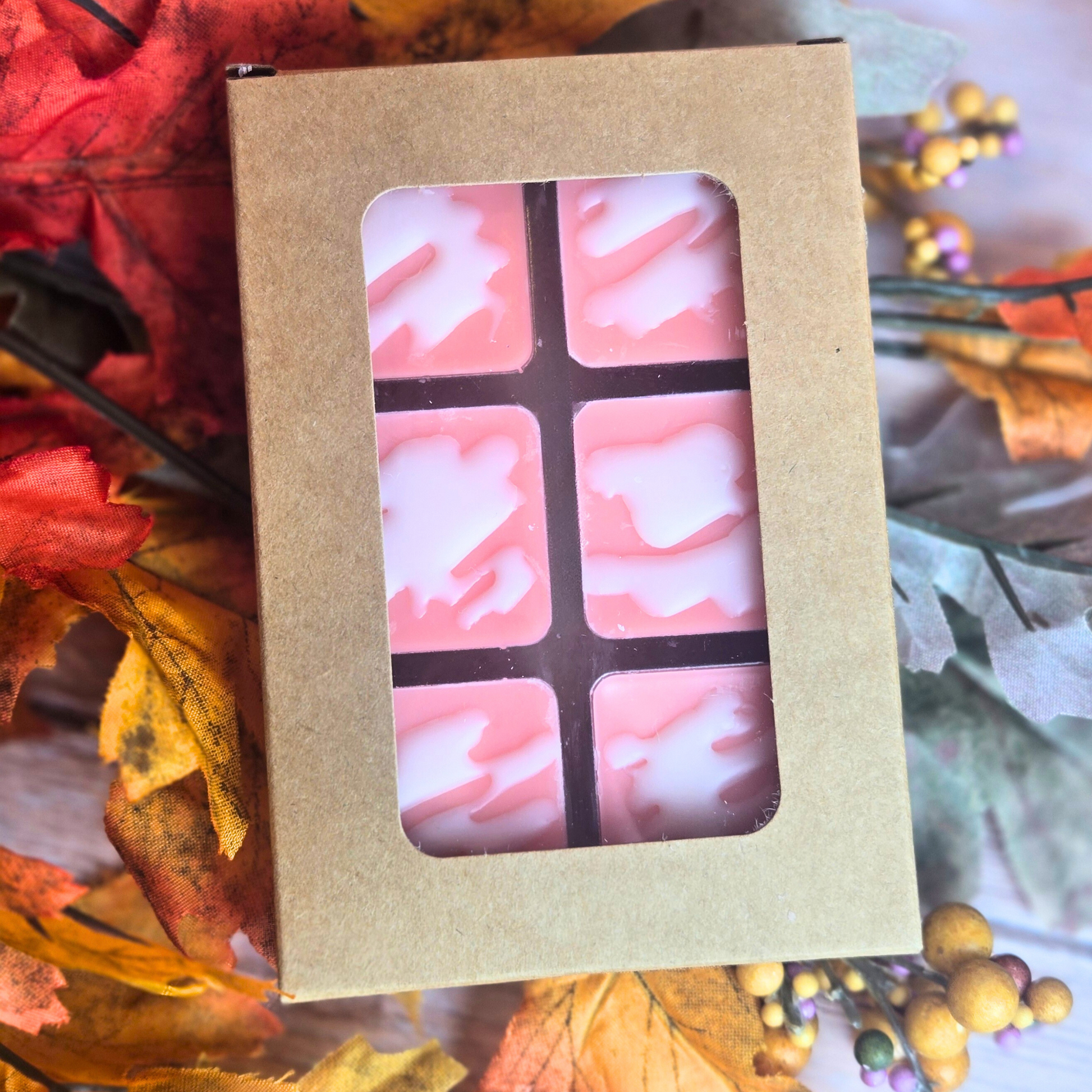 Kraft box of pink and white wax melts sitting on fall leaves, front view.