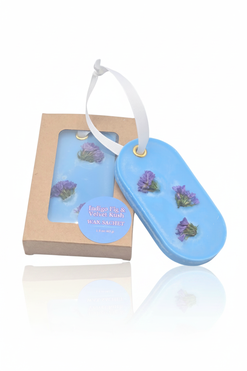 A blue oval wax sachet with three purple dried flowers and a white hanging ribbon, shown next to its kraft paper box labeled 'Indigo Fig & Velvet Wax Wax Sachet'.